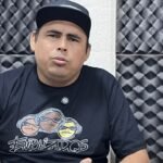 Homem sentado em estúdio acústico, falando com expressão séria, usando boné preto e camisa do coletivo Enraizados, diante de painéis de espuma acústica.