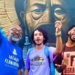 Três artistas do hip-hop posam sorrindo em frente a um mural colorido inspirado em ancestralidade.